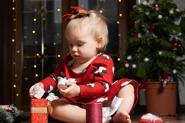 Schönes kleines einjähriges Mädchen mit blonden Haaren, das Weihnachtsgeschenke spielt, Bokeh-Girlanden, Kinder und Silvester