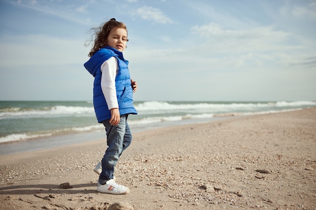 Schönes kleines Kind, das die Kamera durch die Schulter betrachtet, während es am Strand steht. Glückliches Kindheitskonzept. Internationaler Kindertag. Lieben Sie Natur- und Planetenerdeschutzkonzept