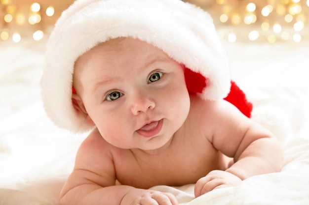 Schönes kleines Kind feiert Weihnachten. Neujahrsfeiertage. Ein Kind in einem Weihnachtskostüm. Kindheits- und Menschenkonzept - glückliches neugeborenes Baby in Weihnachtsmütze über Feiertage beleuchtet Hintergrund.