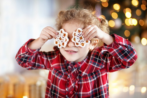 Schönes kleines lächelndes Mädchen, das Weihnachtscoocies über Augen hält. Weihnachtszeit zum Spaß zu Hause. Neujahrsbonbons