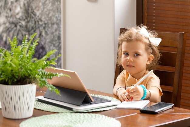 Schönes kleines Mädchen 3 Jahre alt mit lockigem Haar sitzt am Tisch und erhält pädagogische Sprachstunden auf Tablette. frühkindliches Bildungskonzept