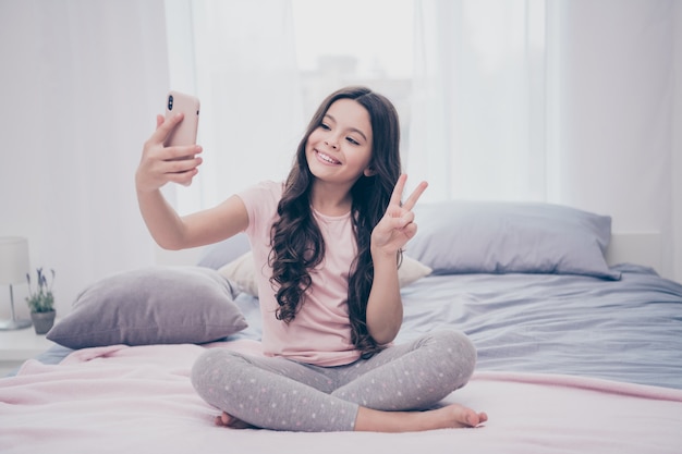 Schönes kleines Mädchen, das auf ihrem Bett mit einem Smartphone sitzt