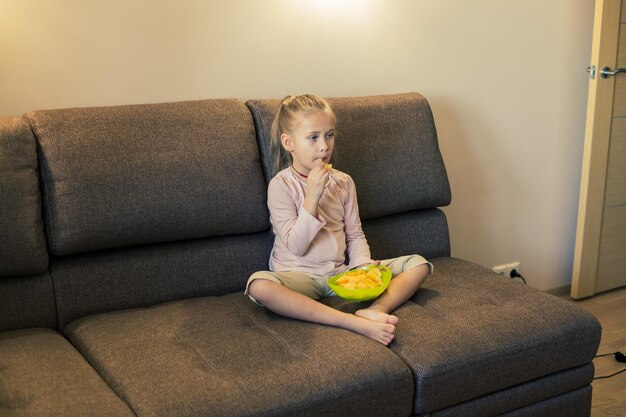 Schönes kleines Mädchen, das beim Fernsehen auf dem Sofa ungesundes Essen isst