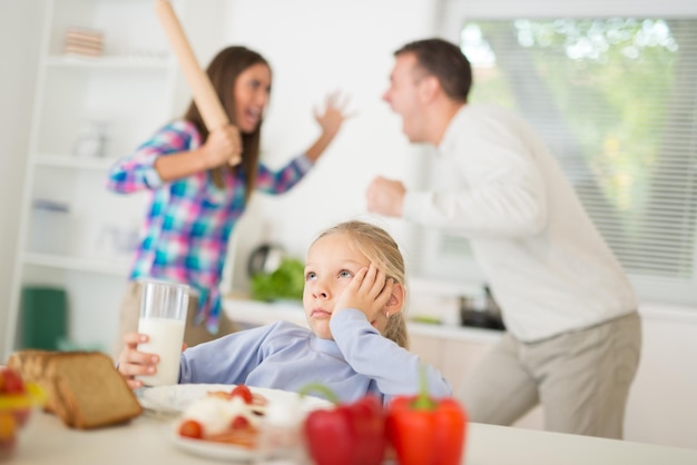 Schönes kleines Mädchen, das in der Küche frühstückt und dem Streit der Eltern zuhört. Sie ist traurig und verärgert, hält den Kopf und schaut weg.