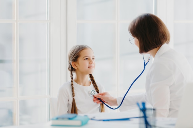 Schönes kleines Mädchen hört Konsultation von professionellen erfahrenen Arzt, der ihre Lungen mit Stethoskop hört