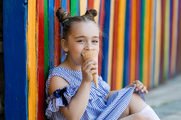 Schönes kleines Mädchen im blauen Kleid, das Eiscreme im bunten Hintergrund des Parks isst