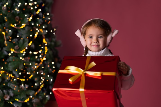 Schönes kleines Mädchen im Hut, das nahe Geschenken und Weihnachtsbaum-Bokeh-Lichtern sitzt