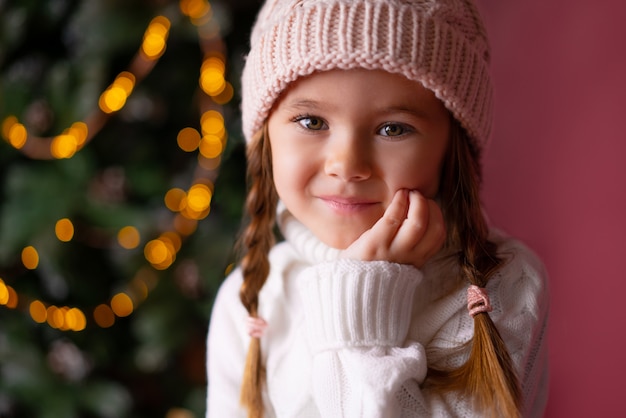 Schönes kleines Mädchen im Hut, der nahe Geschenken und Weihnachtsbaum sitzt