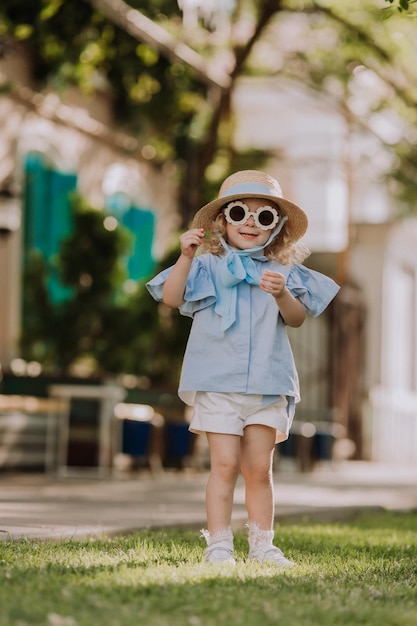 Schönes kleines Mädchen in blauem Kleid, Strohhut und Sonnenbrille, das im Freien spielt, blauer Hintergrund, Karte