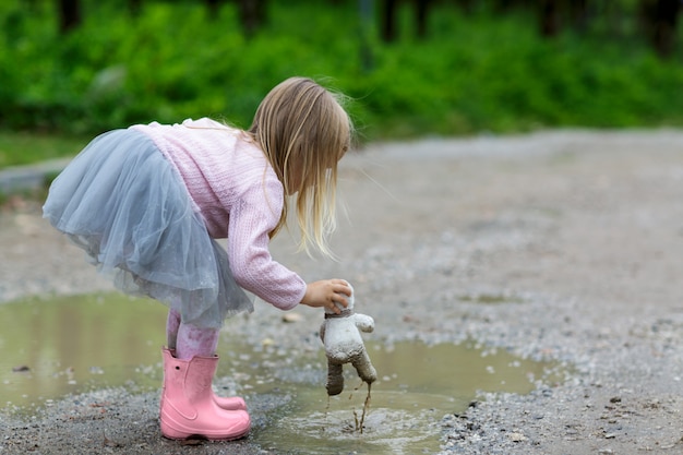 Schönes kleines Mädchen in einem Ballettröckchenrock, der einen Teddybären in einer Pfütze auf der Straße schlägt