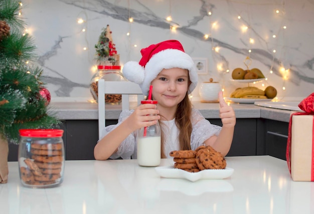 Schönes kleines Mädchen in einer Weihnachtsmütze trinkt Milch und isst am Heiligabend zu Hause Kekse. Das Konzept von Feiertagen, Weihnachten und Kindheit. Selektiver Fokus