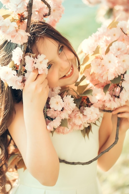 Schönes kleines Mädchen in weißem Glamour-Kleid mit langen Haaren und lächelndem Gesicht im Frühling rosa Blüten blühen sonnigen Tag