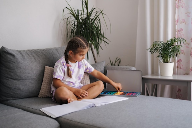 Schönes kleines Mädchen sitzt auf dem Sofa im Wohnzimmer und zeichnet mit farbigen Bleistiften zu Hause Stimmung