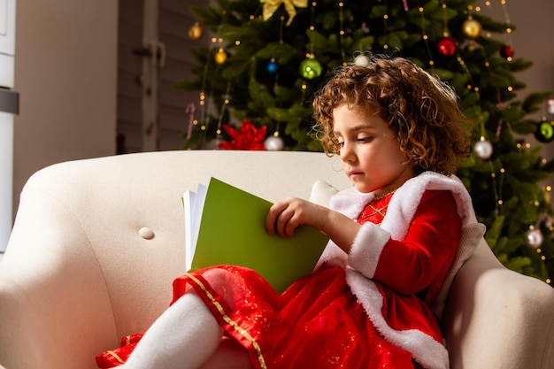 Schönes kleines Mädchen sitzt auf dem Stuhl neben einem schön geschmückten Weihnachtsbaum