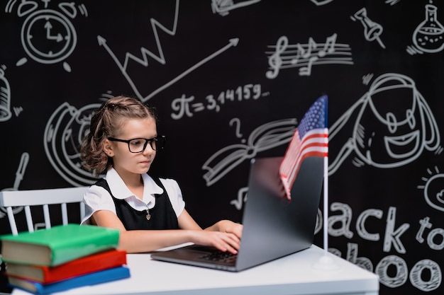 Schönes kleines Schulmädchen am Schreibtisch vor schwarzem Hintergrund mit USA-Flagge