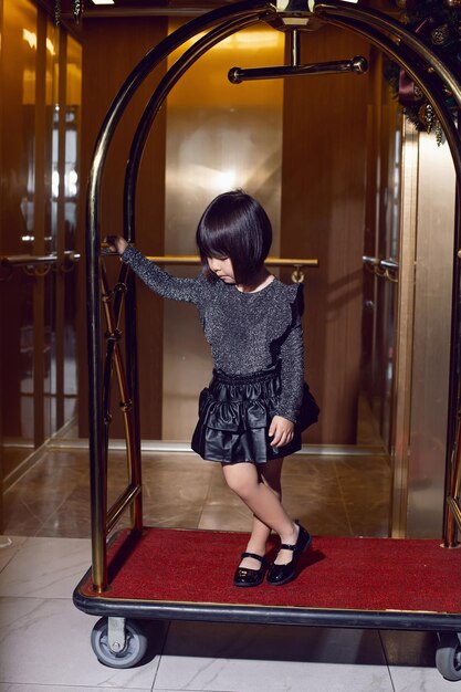 Schönes koreanisches Mädchen in einem schwarzen Kleid sitzt auf einem Gepäckwagen im Hotel in der Nähe des Aufzugs