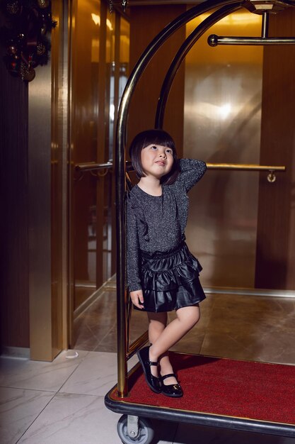 Schönes koreanisches Mädchen in einem schwarzen Kleid sitzt auf einem Gepäckwagen im Hotel in der Nähe des Aufzugs