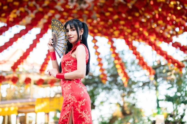 Schönes Lächeln des Porträts Asiatische junge Frau, die rote traditionelle chinesische Cheongsam-Dekoration trägt und ein chinesisches Fanning für das chinesische Neujahrsfest am chinesischen Schrein in Thailand hält