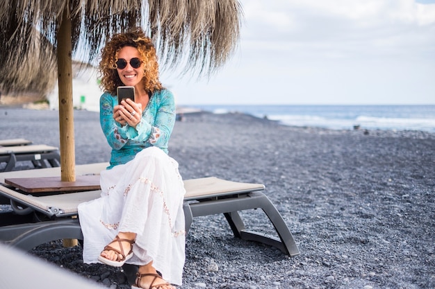 schönes Lächeln schöne einsame Frau mittleren Alters genießen Sie den Strand auf einem Sitz mit Meer sitzen. Verwenden des Smartphones, um sich mit Freunden zu Hause zu verbinden oder mit einem Team-Online-Geschäft zu arbeiten.