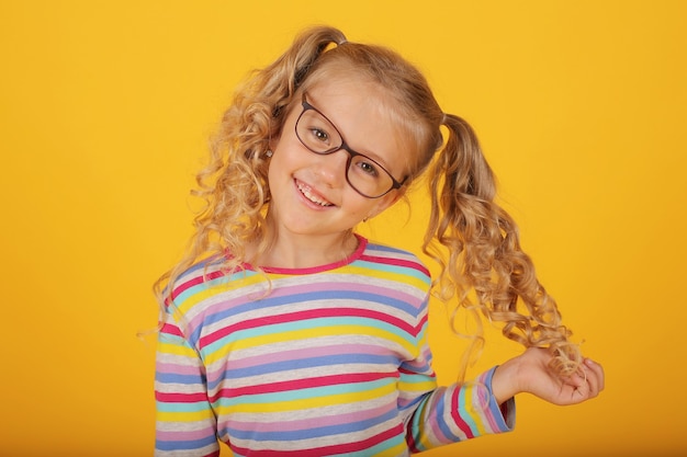 schönes lächelndes blondes Mädchen mit Brille auf gelbem Hintergrund in einer farbigen Jacke Emotionen