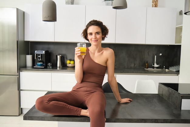 Schönes lächelndes Fitness-Mädchen in Aktivkleidung sitzt auf der Küchentheke und trinkt ein Glas Orangensaft