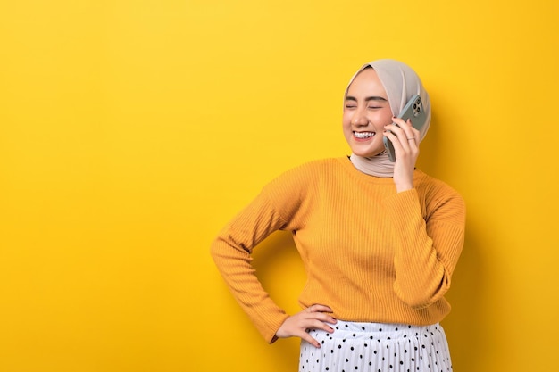 Schönes lächelndes glückliches asiatisches Mädchen, das Hijab trägt und auf dem Handy spricht, mit Kopienraum isoliert auf gelbem Hintergrund