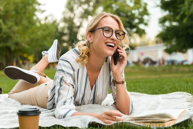 Schönes lächelndes junges blondes Mädchen, das sich auf einem Rasen im Park entspannt und mit dem Handy spricht, während es auf einer Decke liegt