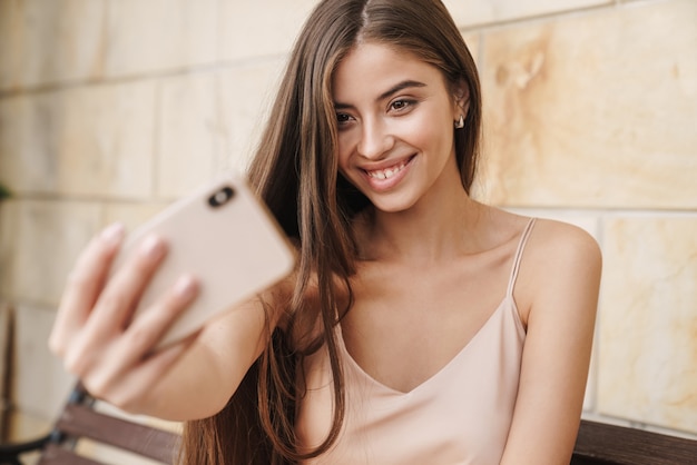 Schönes lächelndes junges Mädchen im Sommerkleid, das draußen auf einer Bank sitzt und ein Selfie macht