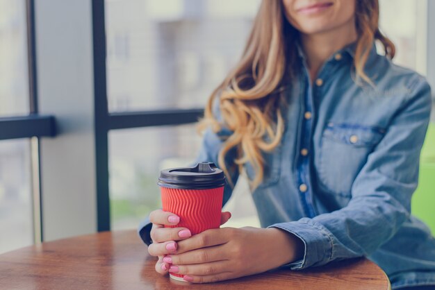 Schönes lächelndes junges Mädchen mit dem lockigen Haar, das im Jeanshemd gekleidet ist, hat eine Pause und trinkt Morgenkaffee zum Mitnehmen