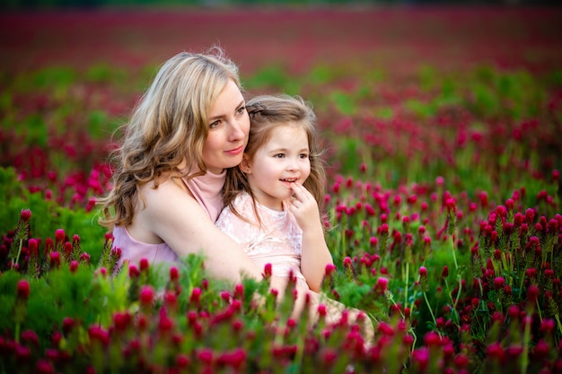 Schönes lächelndes kindermädchen mit junger mutter im familienblick im bereich der kleeblumen in der sonnenuntergangszeit