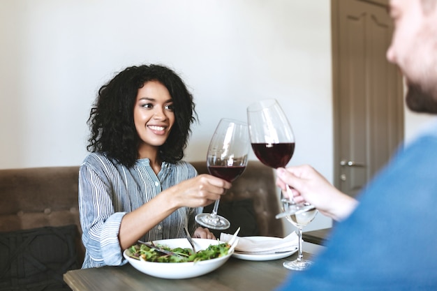 Schönes lächelndes Mädchen, das Rotwein mit Freund im Restaurant trinkt. Hübsches afroamerikanisches Mädchen, das Salat isst und Wein im Café trinkt