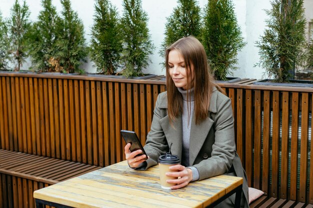 Schönes lächelndes Mädchen mit Brille, das im Café sitzt und Smartphone in den Händen hält und plaudert