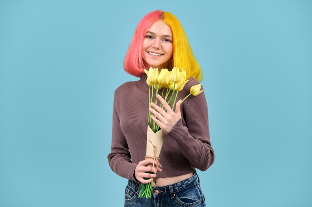 Schönes lächelndes modisches jugendlich Modell mit Blumenstrauß aus gelben Tulpen auf blauem Studiohintergrund. Mädchen mit gefärbten Haaren, im Jeanspullover, Blumen genießend