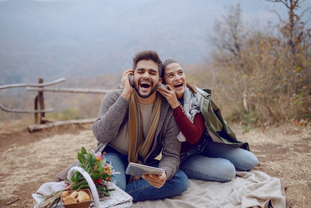 Schönes lächelndes multikulturelles Paar in der Liebe, die auf Decke beim Picknick im Herbst sitzt, mit Tablette und Musik über Kopfhörer hört. Neben ihnen sind Hund und Korb mit Futter.