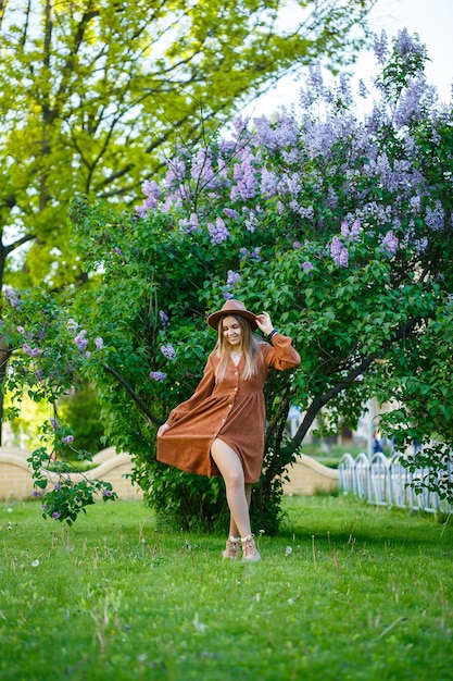 Schönes langhaariges Mädchen in einem Hut auf einem Hintergrund von lila Blumen. Sie hat einen europäischen Auftritt mit einem Lächeln im Gesicht. Junge Frau, die an einem warmen sonnigen Tag in einem blühenden Park geht