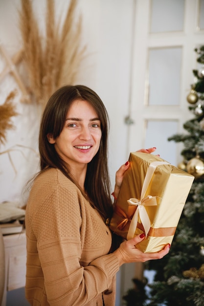 Schönes langhaariges Mädchen nahe Weihnachtsbaum, das goldenes Geschenk hält und in die Kamera lächelt
