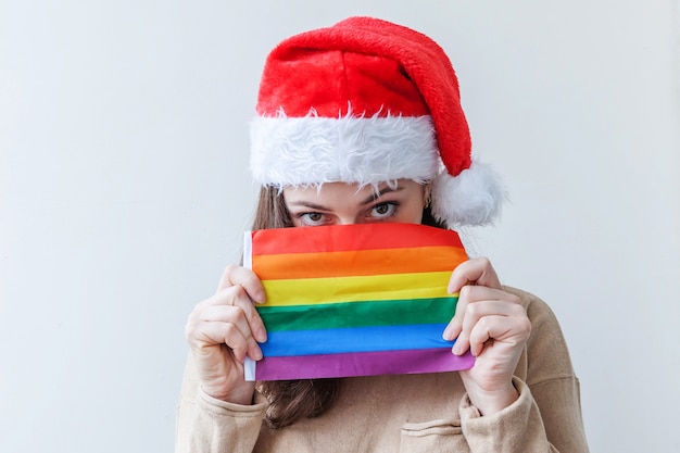 Schönes lesbisches Mädchen im roten Weihnachtsmannhut mit LGBT-Regenbogenfahne lokalisiert auf weißem Hintergrund