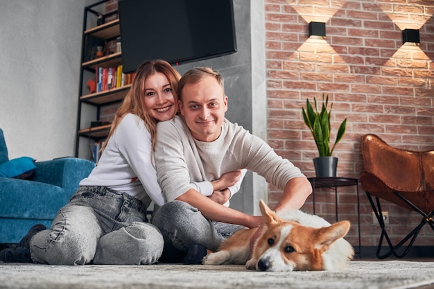 Schönes liebevolles Paar, das mit süßem Corgi auf dem Boden sitzt