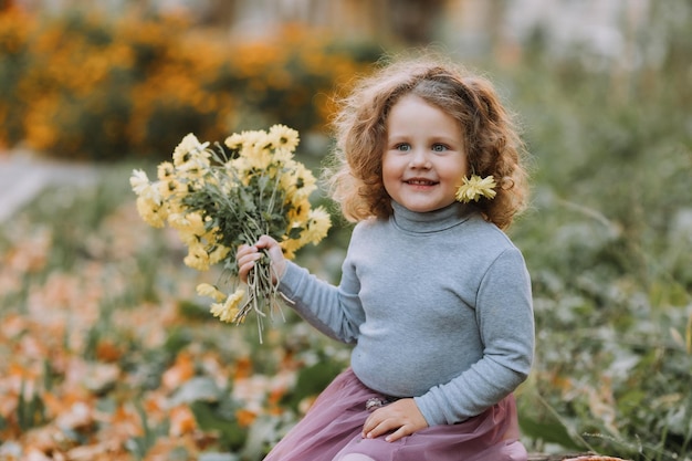 schönes lockiges kleines Mädchen im blauen Hemd im Park mit Blumen zur Herbstzeit Herbstgesundheitskarte