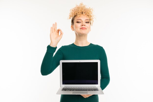 Schönes lockiges Mädchen, das einen Laptop mit Modell in ihren Händen auf weißer Wand hält