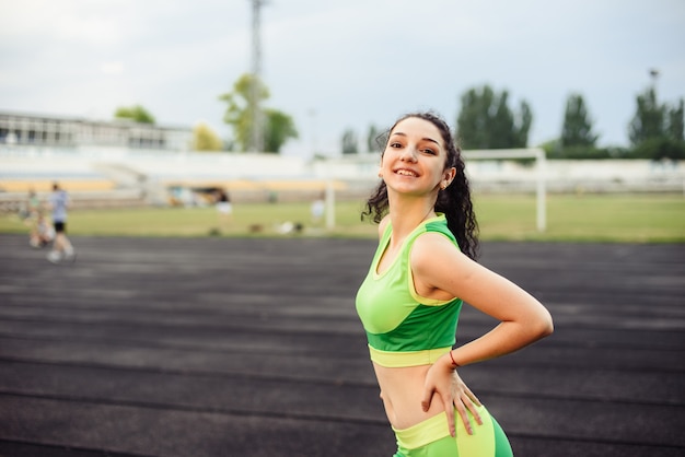 Schönes lockiges Mädchen, das Muskeln im Stadion biegt. Das Mädchen macht Sport. Hellgrüner Trainingsanzug