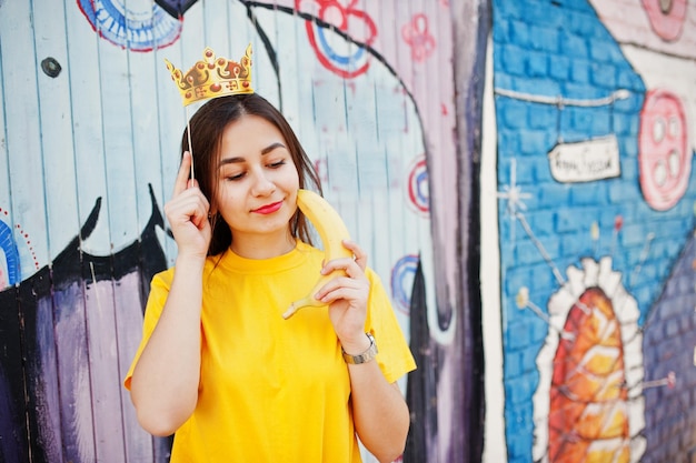 Schönes, lustiges Teenager-Mädchen mit Banane in der Hand trägt eine gelbe T-Shirt-Krone auf einem Stock in der Nähe einer Graffiti-Wand