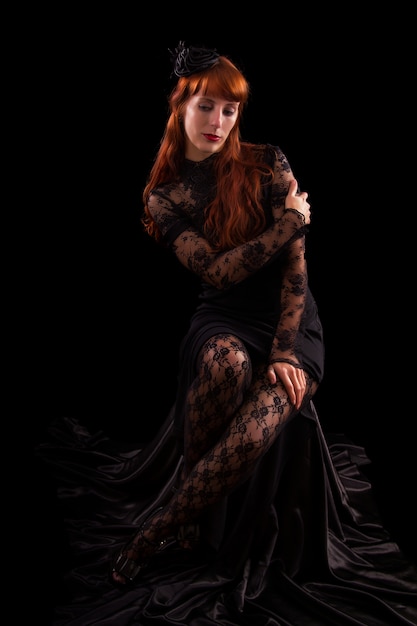 Schönes Mädchen auf einem schwarzen gotischen Kleid mit dem roten Haar lokalisiert auf einem schwarzen Hintergrund.