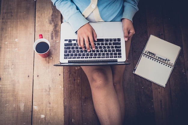 Schönes Mädchen benutzte Laptop und Kaffeetasse in den Händen des Mädchens, die auf einem Bretterboden sitzen