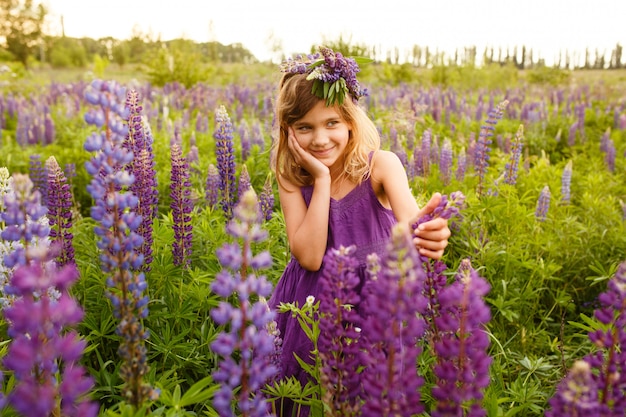 Schönes Mädchen, das in einem violetten Kleid mit einem Kranz von Lupinen lächelt