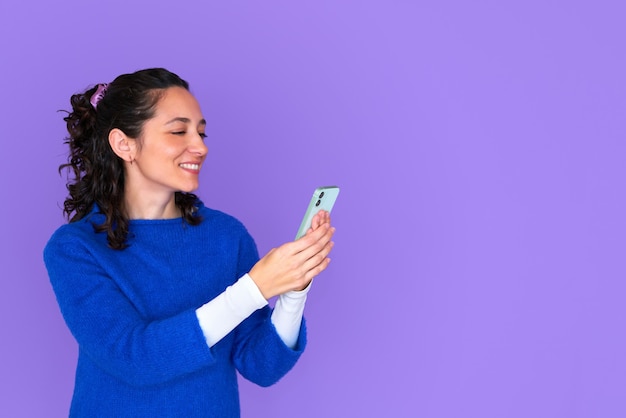 Schönes Mädchen, das selfie auf lokalisiertem lila Hintergrund trägt, der blauen Pullover trägt. Smartphone mit einer Hand halten, zur Kamera lächeln. Lockige haare.