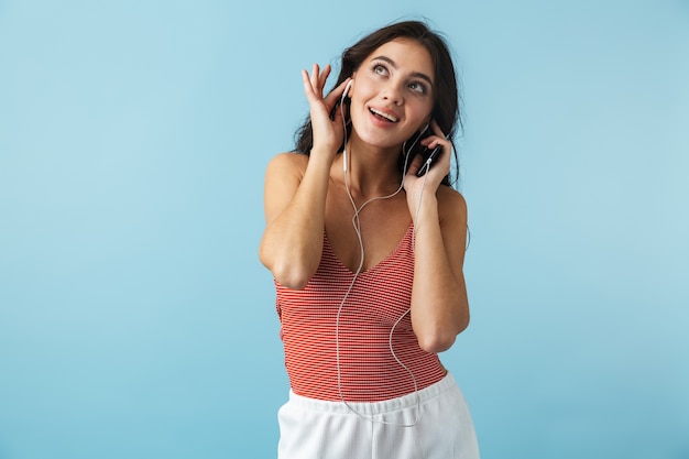 Schönes Mädchen, das Sommerkleidung trägt, die lokal über Blau steht, Musik mit Kopfhörern hörend, Handy haltend