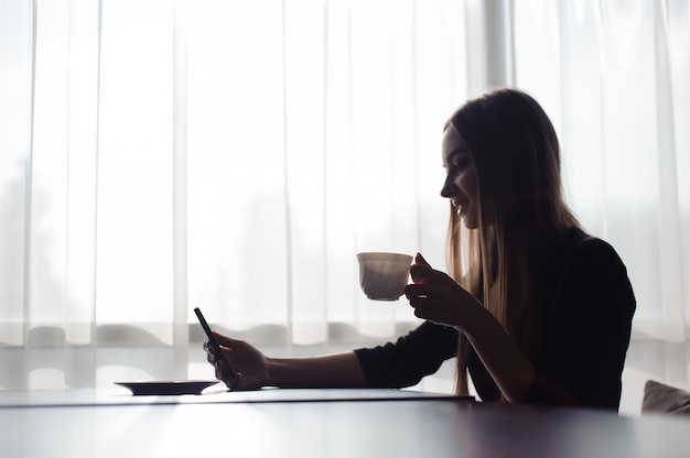 Schönes Mädchen, das Tee trinkt und ein Handy benutzt, während es in einem Café vor einem großen Fenster sitzt