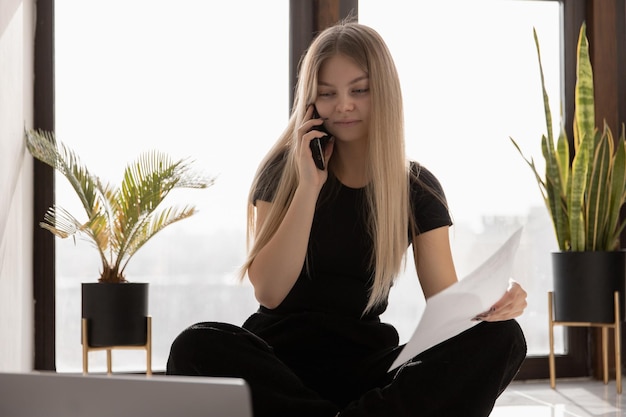 Schönes Mädchen, das zu Hause am Computer arbeitet, sitzt auf dem Boden am Fenster und telefoniert lächelnd