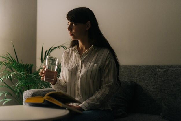 Schönes Mädchen hält ein Glas Wasser in einem dunklen Raum. Auf dem Tisch liegt ein Buch. Frau schaut weg.